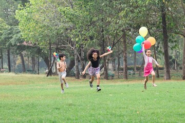 Group of elementary diverse children, boy and girls running joyfully through a park holding balloons and pinwheels. African-American girl leading, reflecting happiness excitement, and carefree in park