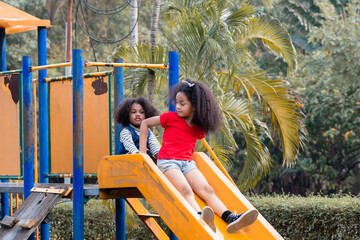 Two African American sibling girls joyful and laughing sliding down a yellow playground together,...