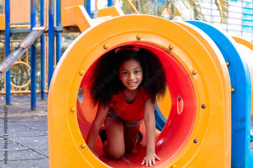 Wall mural young afro hair elementary schoolgirl crawling through colorful tunnel on a playground, weekend care