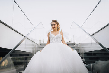 Beautiful young curly smiling blonde bride in white long dress walking down stairs in city...