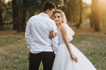 Fashionable groom and beautiful smiling bride curly blonde in white long dress stand hugging outdoors in forest in nature at sunset. Wedding photography, portrait of happy newlyweds in love.