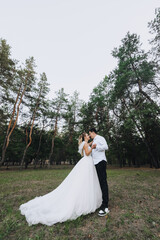Young groom and beautiful smiling bride curly blonde in white lace long dress standing hugging outdoors in forest in nature at sunset. Wedding photography, portrait of happy newlyweds in love.