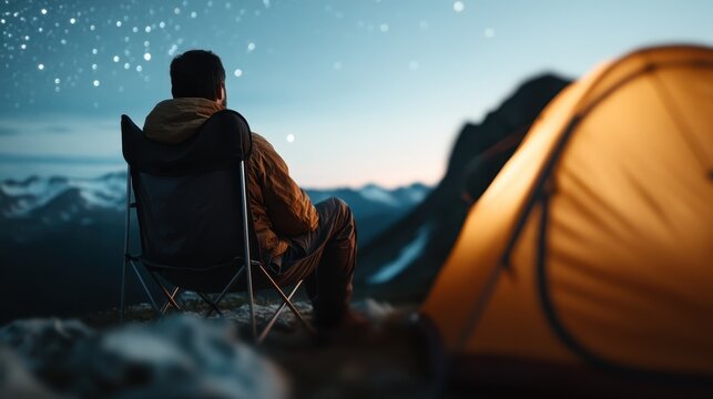 A person sits on a camping chair, gazing at the night sky filled with stars, next to an illuminated tent on a mountain with snow-capped peaks in the background.