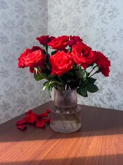 A vibrant bouquet of red roses in a textured glass vase is captured on a wooden table, illuminated by soft natural sunlight. The scattered rose petals on the surface add a touch of romance to the