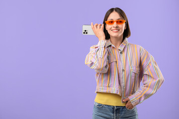 Beautiful woman in sunglasses with smartphone listening to voice message on violet background,...