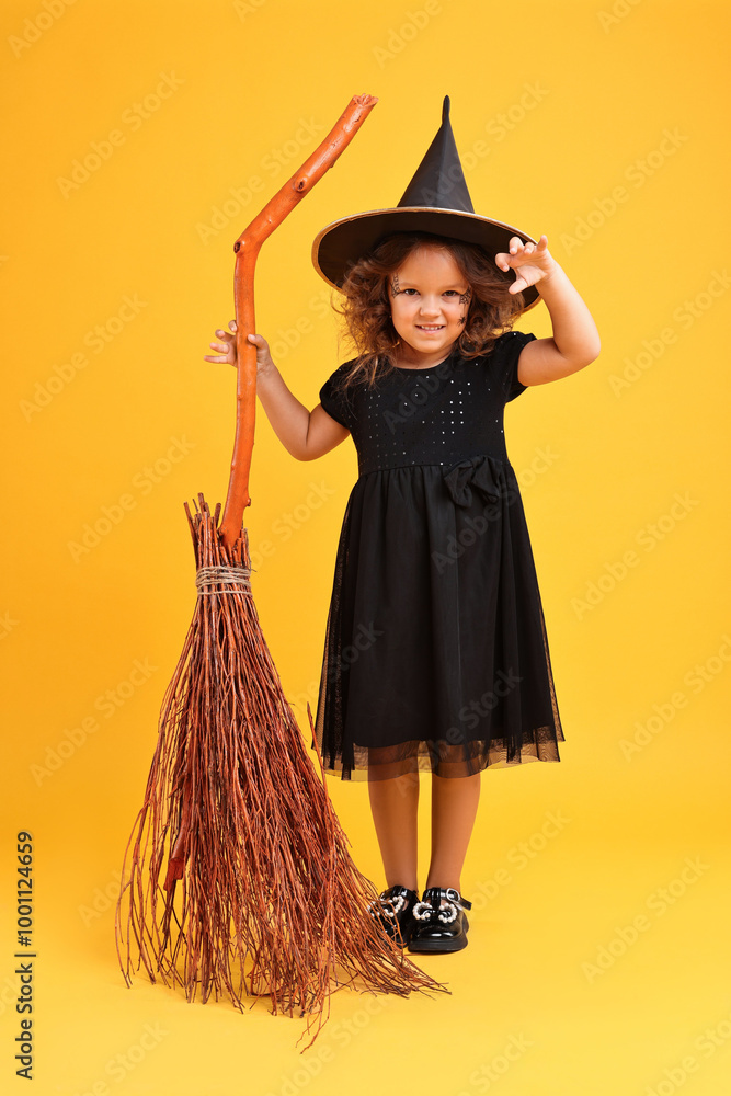 Wall mural Funny girl with broom dressed like witch on yellow background. Halloween celebration