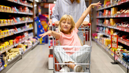 Little girl having fun riding in a shopping cart while shopping groceries at the supermarket with her mother