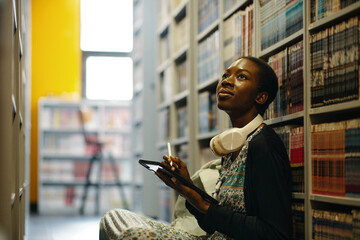 African American woman using digital tablet while sitting on the floor among bookshelves in the library