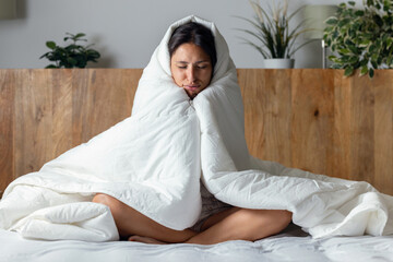 Tired and cold woman covered with the duvet sitting on the bed at home