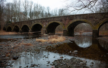 An ancient moss-covered stone arch bridge crosses a quiet river surrounded by bare trees in winter, in a serene natural setting, Pennsylvania, USA
