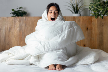 Tired and cold woman covered with the duvet sitting on the bed at home