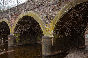 An ancient moss-covered stone arch bridge crosses a quiet river surrounded by bare trees in winter, in a serene natural setting, Pennsylvania, USA