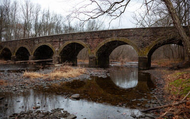 An ancient moss-covered stone arch bridge crosses a quiet river surrounded by bare trees in winter, in a serene natural setting, Pennsylvania, USA