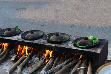 A traditional Indonesian serabi-making process, showing hands pouring batter onto a clay pan over a wood-fired stove. The smoky atmosphere and authentic tools emphasize the local street food culture.