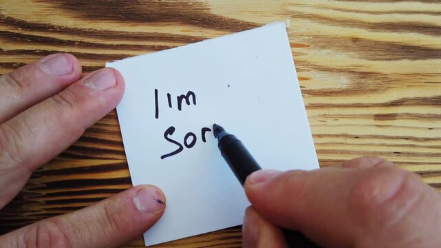 A man writes the phrase I'm sorry on a white sticky note on a table background