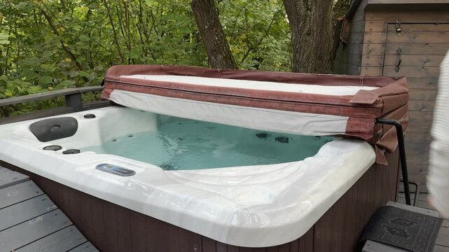 A man in a robe lifts the lid of a bubbling outdoor heated Jacuzzi, revealing the inviting water inside. The scene conveys the anticipation of a pleasant evening of relaxation in the fresh air.