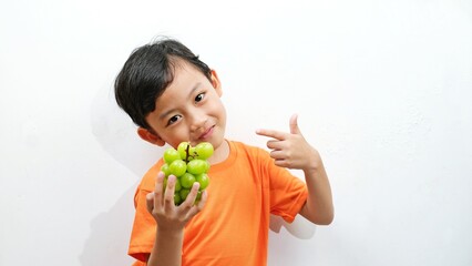 A young boy in an orange shirt holds a bunch of fresh green Shine Muscat grapes, smiling and pointing at them. Healthy, nutritious, and sweet, these grapes are perfect for promoting healthy eating