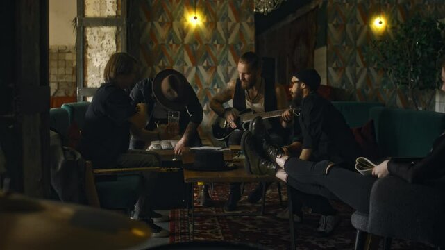 Full shot of diverse music group members greeting male guitarist wearing hat while sitting in bar at weekend, relaxing and having drinks before concer