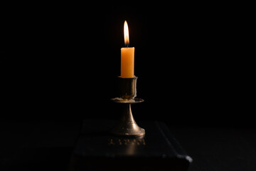 Dark, an antique candle cast its light on the old Bible, its flames flickering gently over the book on the table, symbolizing the enduring essence of religion. candle, bible, dark, light, book, old.