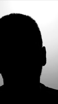 Young caucasian men black silhouette against grey wall talking during interview, hidden identity. Secret testimony of crown witness. The seizure victim answers questions. Prisoner of war interview.