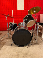 Classic black drum set is positioned in a studio with vibrant red soundproof walls. Shiny black drums and bright red background creates a dynamic and energetic music atmosphere. Musical instrument and