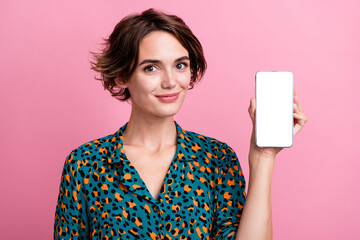 Photo of optimistic girl dressed stylish shirt presenting ui design on smartphone screen empty space isolated on pink color background