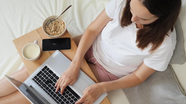 Top view of unrecognizable woman typing on laptop while comfortably working from bed engaging in online learning and networking through social media and email in cozy bedroom