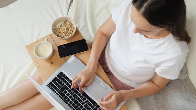 Unrecognizable female journalist typing on laptop while browsing web and writing article on e-commerce connected online through social media and comfortably working from bed