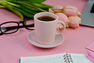 Flatlay of coffee with pink office tools, pink headphone, notebook, pen and a bouquet of pink tulips on pink background as copy space concept