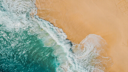 Waves from the turquoise ocean gently crash onto the golden sands of Kelingking Beach, Nusa Penida, Indonesia. The aerial view showcases the stunning contrast between the sea and the shore.