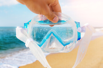 A man holds a snorkel mask in his hand against a clear blue sky and turquoise ocean. Diving mask in a man's hand. Photo of a mask and snorkel for swimming in a pool or sea with a central composition
