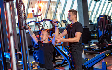 Men training together at a gym during sunset. A personal trainer assists a man performing lat...