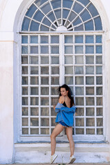 Woman in denim jacket poses by large window. A young woman stands confidently by a large, arched window wearing a denim jacket and shorts, enjoying the warm sunlight.