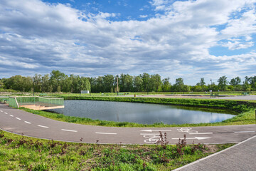 Serene park scene with bike path alongside calm pond. Small wooden deck extends over water.