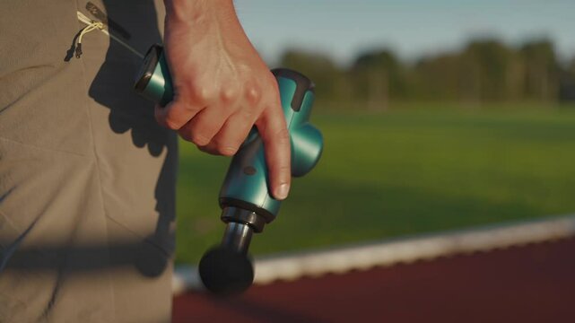 A male athlete massages muscles and tendons with a massage percussion device after a workout at the stadium. Sportsman uses electric massager gun in hand massaging the muscle. Sports recovery concept