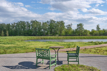 Serene outdoor scene in park. Two green wooden chairs, round table. Calm pond, surrounded by trees.