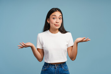 Confused young Asian woman shrugging with raised arms and open palms on blue background