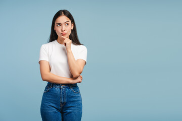 Pensive young Asian woman thinking with finger on chin posing on blue background copy space