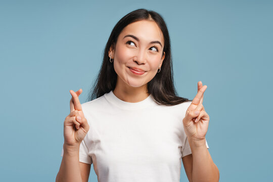 Young, smiling Asian woman crossing fingers making wishful expression on blue background