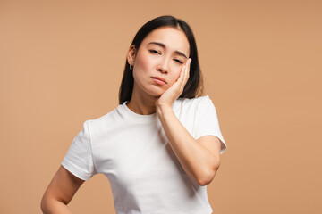 Asian woman suffering from strong toothache touching cheek, looking at camera on beige background