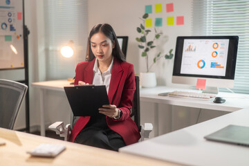 A woman in a red jacket is sitting at a desk with a clipboard in front of her