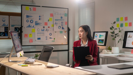 A woman in a red jacket is sitting at a desk with a clipboard in front of her