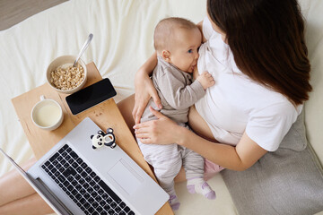 Young mom embracing infant and browsing data on laptop sitting on bed and working on remote project at home while breastfeeding her infant son or daughter