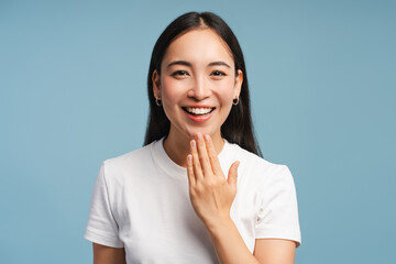 Smiling asian woman is laughing and covering her mouth with hand closeup isolated on blue background