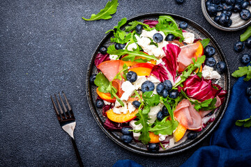 Foodie food salad with peach, jamon, soft cheese, blueberries and arugula on black background, top view