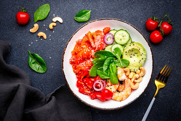 Seafood salad bowl with salmon, shrimp, avocado, spinach, cucumber, tomato, cashew and sesame. Black background, top view
