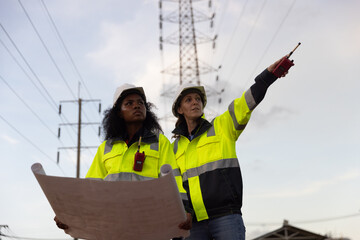 Diverse two women electrical engineers holding blueprints looking confident with power line towers to project planning work producing electrical energy high voltage, energy concept.