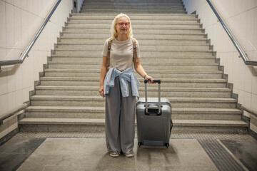 A blonde woman with glasses and a suitcase stands poised at the bottom of the stairs in a subway...