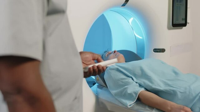 Mature Caucasian woman wearing blue gown lying on CT scanner bed having screening procedure, unrecognizable doctor using controller