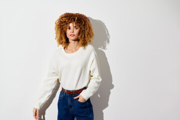 Woman with curly hair in a white sweater and blue jeans, posing against a neutral background, showcasing a casual and stylish look with soft shadows